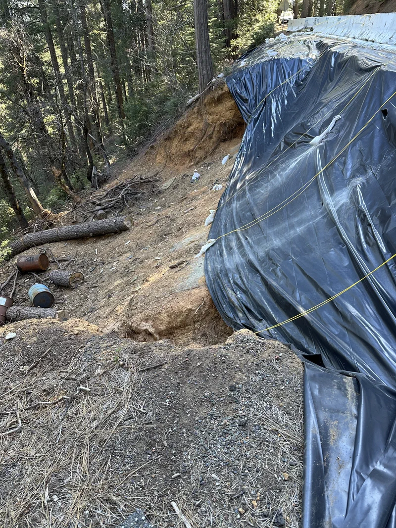 Looking down the failed slope below the roadway, where plastic sheeting is draped over raw soil and fallen timber lies at the bottom