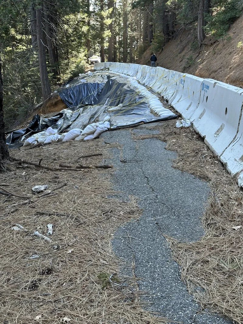 The narrow strip of pavement left at the failure, running between the tarped and sandbagged slope on one side and concrete K-rail on the other